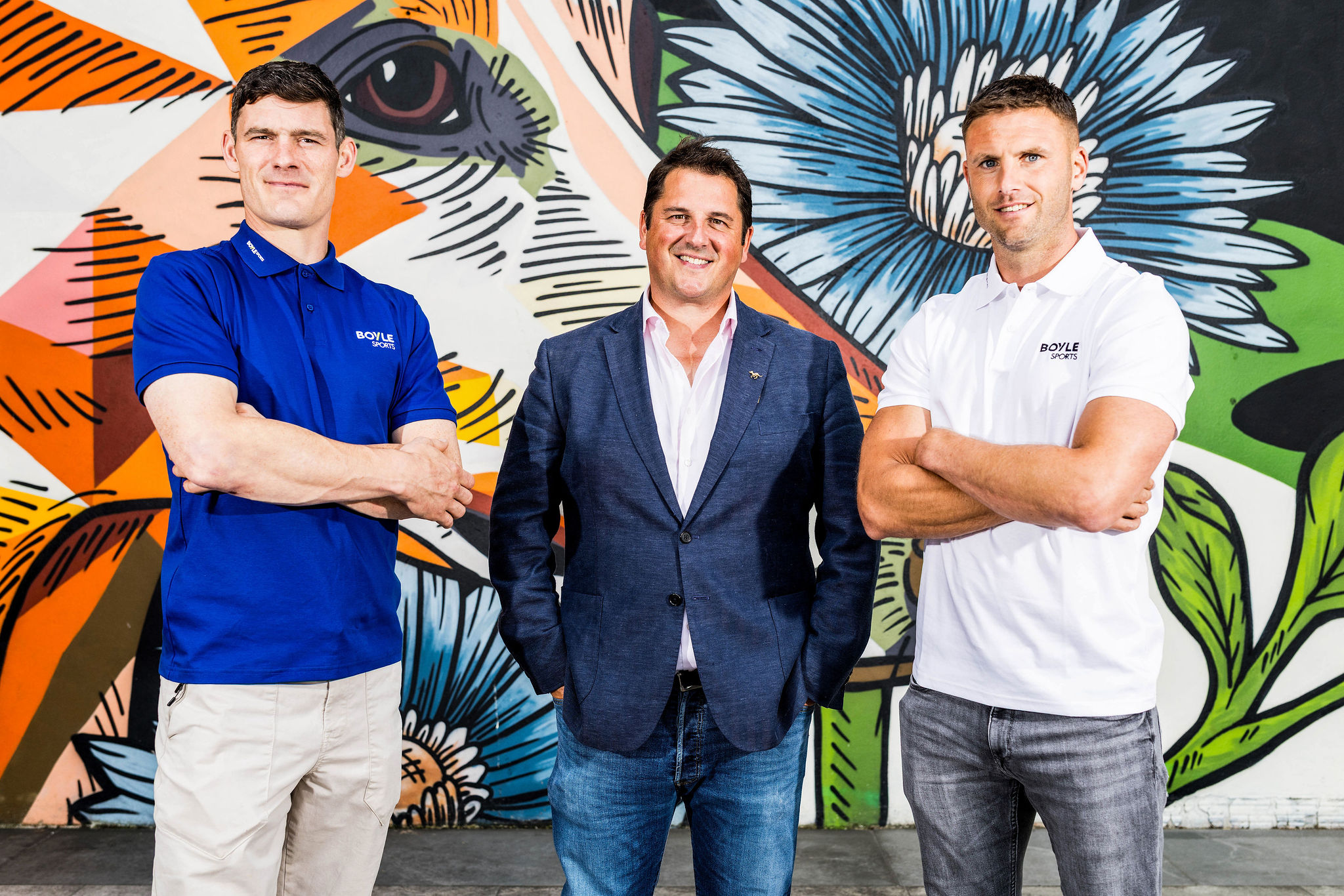 The image shows 3 men, Diarmuid Connolly, Ian Fortune and Eoin Cadogan, against a colourful mural of a greyhound outside Dublin's Shelbourne Park Greyhound Stadium to launch their Celebrity Commentator Challenge