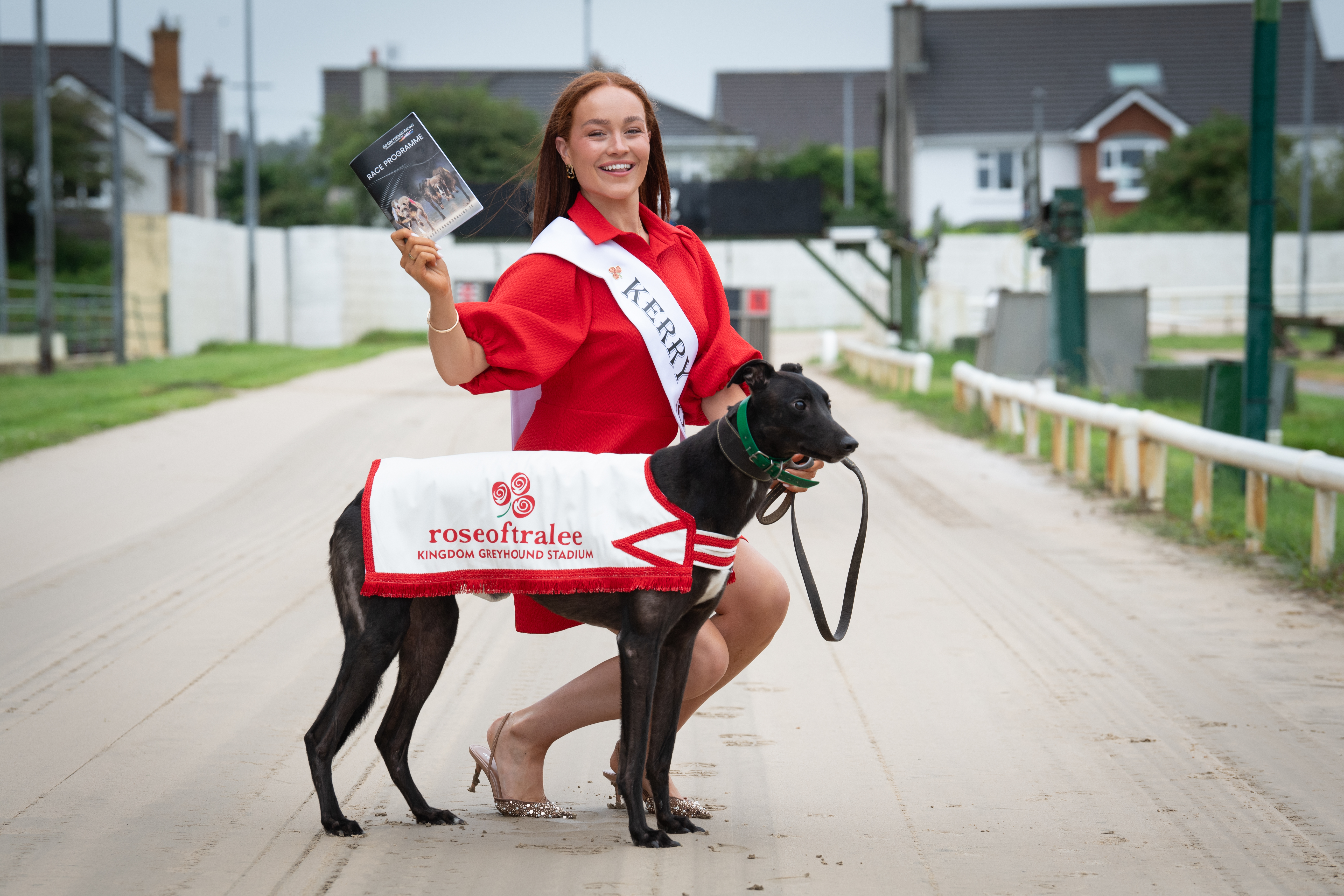 A woman in a red dress, the 2025 Kerry Rose Laura Daly, is pictured on the sand track at the Kingdom Greyhound Stadium in Tralee with a black greyhound wearing a Rose of Tralee branded jacket