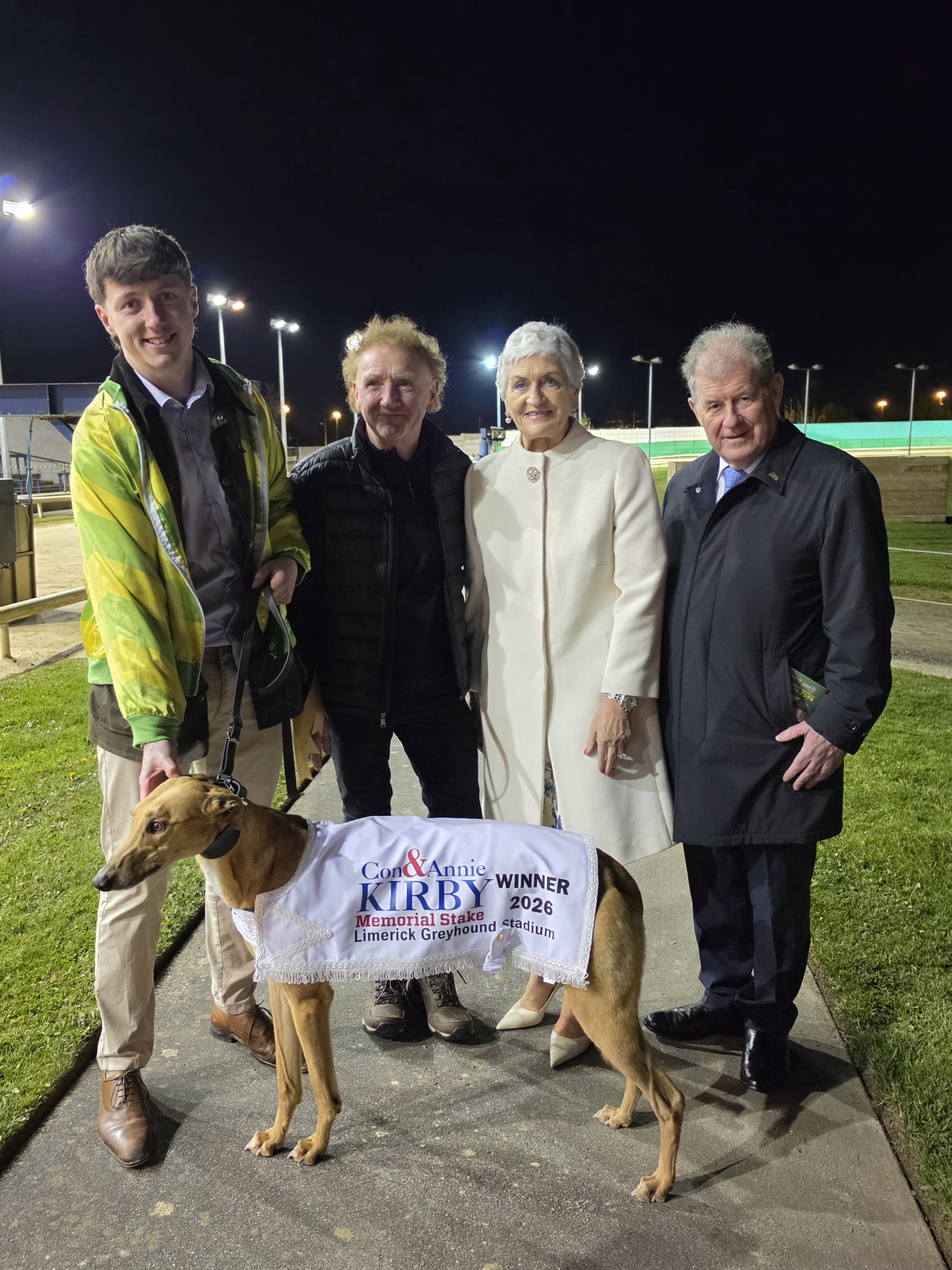 Image shows a fawn coloured greyhound, Bouncing Monarch, wearing a winner's jacket for the Con and Annie Kirby Memorial, standing in front of 4 people, Mark O'Donovan, Michael O'Donovan, Noreen McManus and JP McManus at Limerick Greyhound Stadium with the track and stadium behind them and a dark night sky